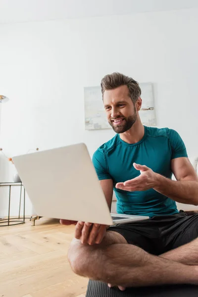 Lächelnder Mann zeigt auf Laptop, während er in Lotus-Pose auf Yogamatte sitzt — Stockfoto