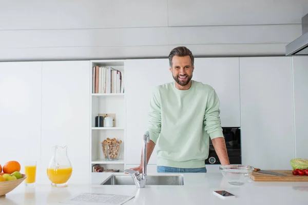 Uomo allegro sorridente a macchina fotografica vicino a succo d'arancia e lavandino in cucina — Foto stock