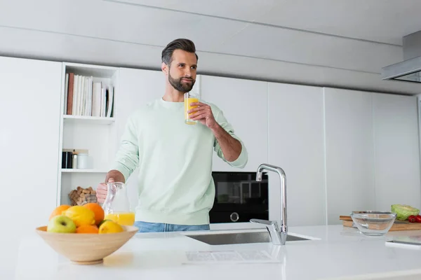 Pleased man drinking orange juice near fresh fruits in kitchen — Stock Photo