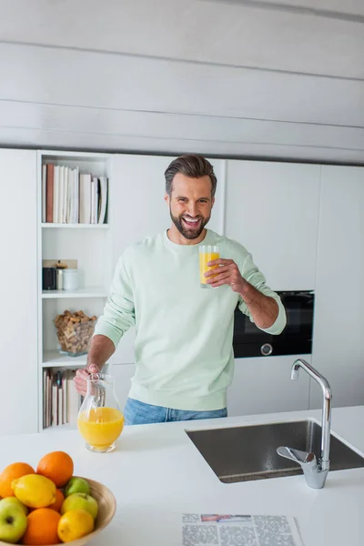 Смеющийся мужчина держит стакан свежего апельсинового сока на кухне — стоковое фото