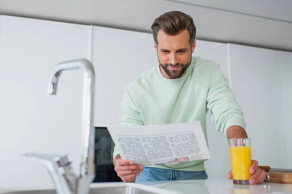 Hombre positivo leyendo periódico cerca de vaso de jugo de naranja y grifo en primer plano borroso - foto de stock
