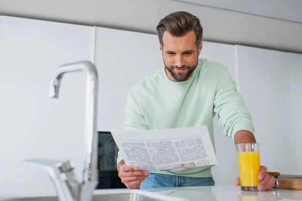 Positiver Mann liest Zeitung in der Nähe von Orangensaft und Wasserhahn auf verschwommenem Vordergrund — Stockfoto