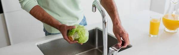 Частичный вид человека, держащего свежий салат рядом с краном и раковиной на кухне, баннер — стоковое фото