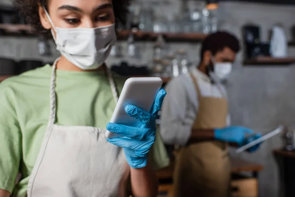 Smartphone in hand of african american barista in medical mask on blurred background — Stock Photo