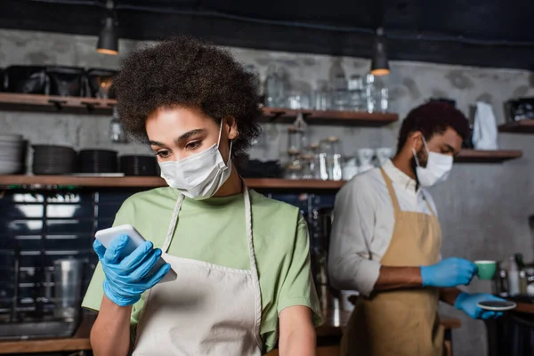 Barista afroamericano in maschera medica e guanto di lattice utilizzando smartphone vicino collega in caffè — Foto stock