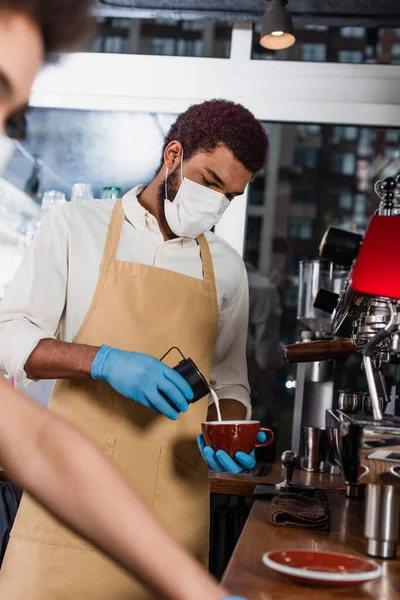 African american barista in medical mask and latex gloves making coffee near blurred colleague in cafe — Stock Photo