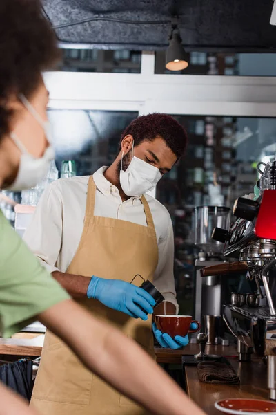 African american barista in protective mask and latex gloves making coffee near blurred colleague — Stock Photo