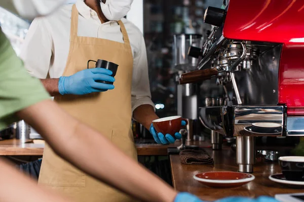 Vista cortada do barista afro-americano em máscara médica e luvas de látex segurando xícara e fazendo café perto colega borrado — Fotografia de Stock