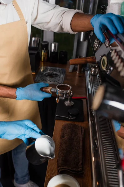 Cropped view of african american barista in latex gloves holding coffee in holder near coffee machine and colleague — Stock Photo