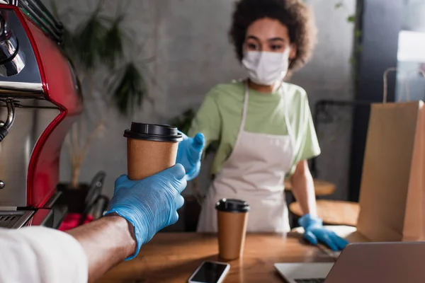 Coffee in hand of african american barista in latex glove and colleague on blurred background — Stock Photo