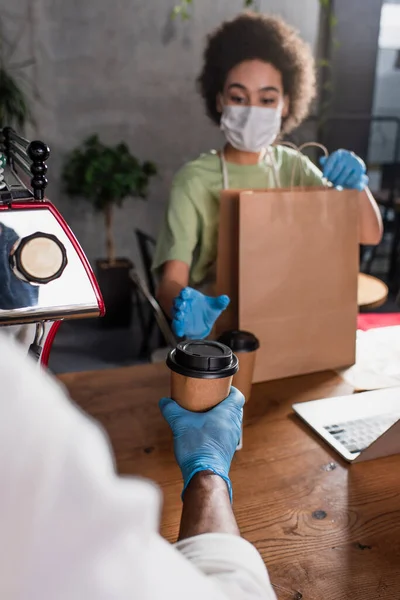 African american barista in latex glove giving coffee to blurred colleague with paper bag in cafe — Stock Photo
