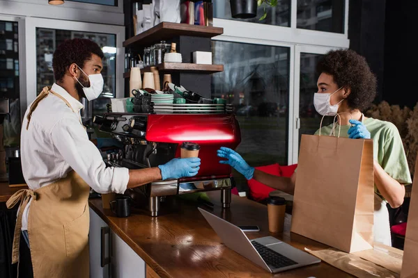 Baristas afro-américaines en masques médicaux et gants en latex tenant du café à emporter et sac en papier dans un café — Photo de stock