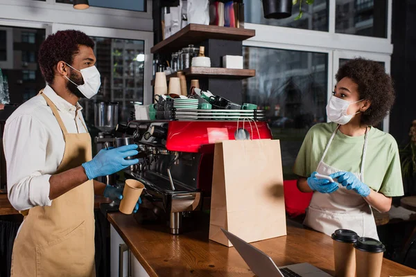 Африканская американская бариста в медицинской маске и латексных перчатках держит кофе, чтобы подойти к коллеге со смартфоном и бумажной чашкой — стоковое фото