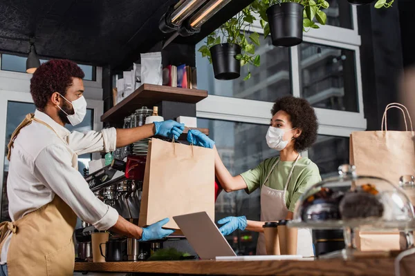 African american barista in medical mask giving paper bag to colleague near coffee to go and laptop — Stock Photo