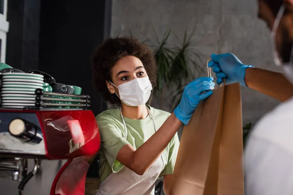 Blurred african american barista in latex glove giving paper bag to colleague in medical mask — Stock Photo