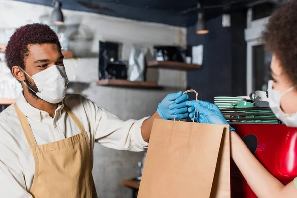 Barista afroamericano in maschera medica che dà sacchetto di carta al collega offuscato vicino alla macchina del caffè — Foto stock