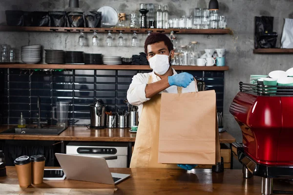 African american barista in latex gloves and medical mask holding paper bag near devices in cafe — Stock Photo