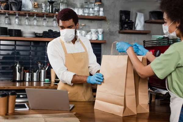 Afroamericano barista in maschera medica dando sacchetti di carta al collega vicino laptop in caffè — Foto stock