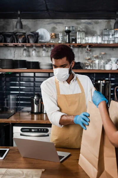 African american barista in medical mask and latex glove looking at laptop and giving paper bags to colleague — Stock Photo