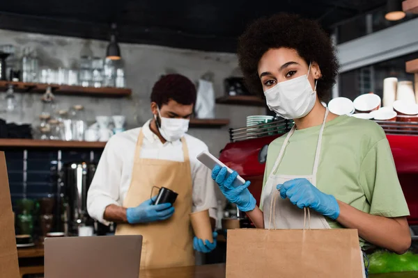 Africano americano barista em máscara médica e luvas de látex usando smartphone e segurando saco de papel perto colega — Fotografia de Stock