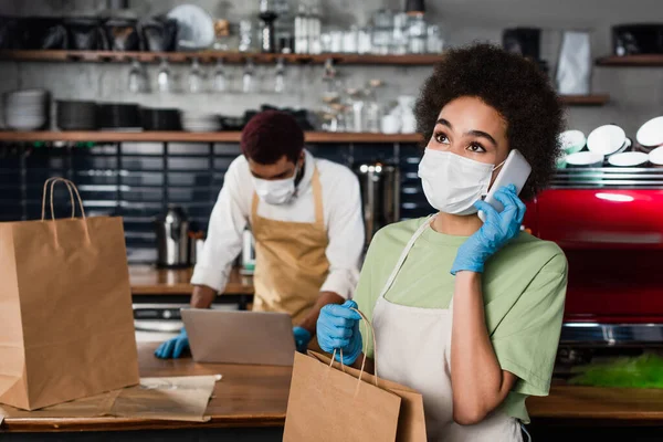 African american barista in medical mask talking on smartphone and holding paper bag in cafe — Stock Photo