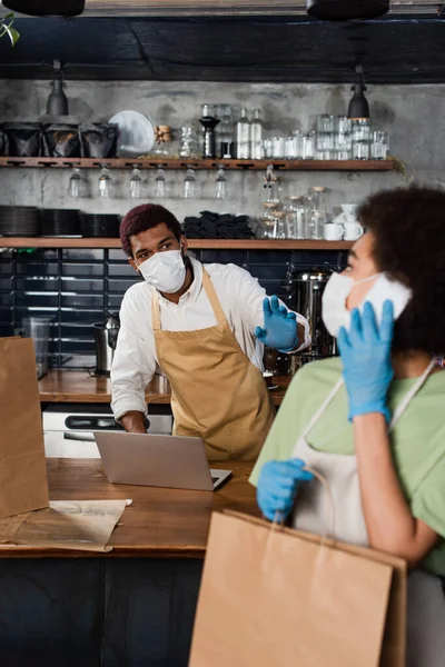 African american barista in medical mask waving hand near blurred colleague with paper bag talking on smartphone — Stock Photo