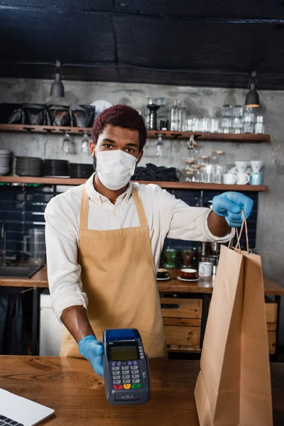 Barista afro-américaine en masque médical et gants en latex tenant terminal de paiement et sac en papier — Photo de stock