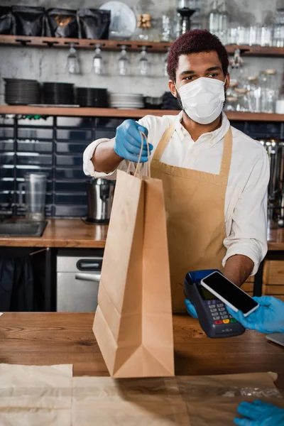 Barista afroamericano en máscara médica sosteniendo bolsa de papel y terminal de pago cerca del cliente con teléfono inteligente - foto de stock