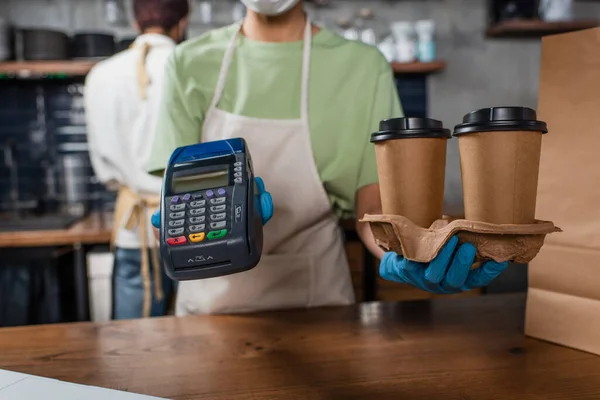 Ausgeschnittene Ansicht eines afrikanisch-amerikanischen Baristas in Latexhandschuhen mit Kaffee zum Mitnehmen und Zahlungsterminal — Stockfoto