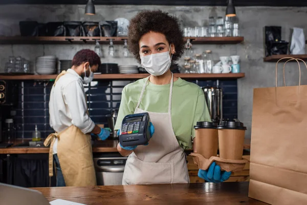 Barista afroamericano en máscara médica y guantes de látex sosteniendo vasos de papel y terminal de pago - foto de stock