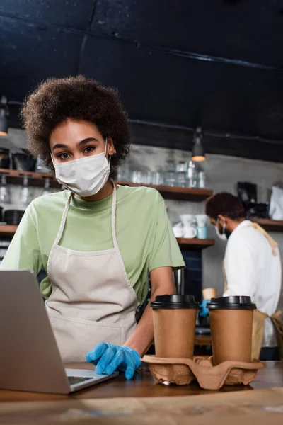 African american barista in medical mask and latex glove using laptop near coffee in cafe — Stock Photo