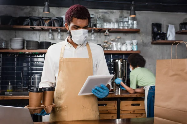 Africano americano barista em máscara médica segurando café para ir e usando tablet digital — Fotografia de Stock