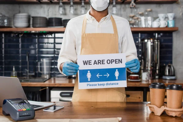 Vista recortada de barista afroamericano en máscara médica y guantes de látex sosteniendo letrero con estamos letras abiertas en la cafetería - foto de stock