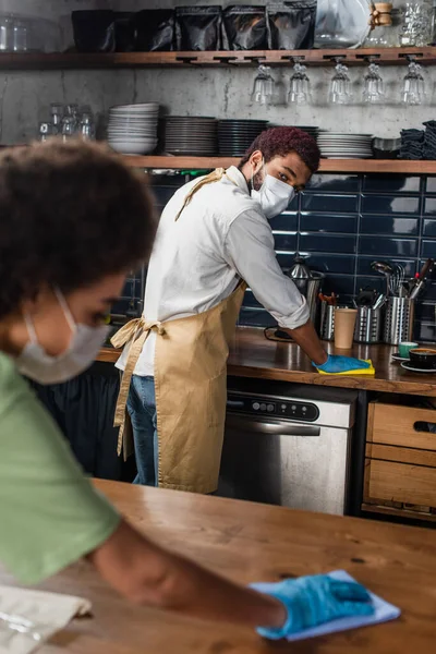 African american barista in medical mask and latex glove cleaning worktop near blurred colleague — Stock Photo