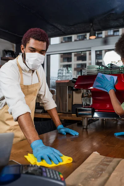 African american barista in medical mask cleaning bar near colleague in cafe — Stock Photo