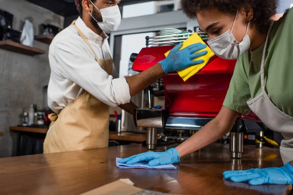 Africano americano barista em máscara médica e luvas de látex bar de limpeza perto colega e máquina de café — Fotografia de Stock