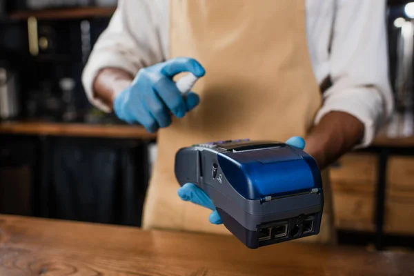 Cropped view of payment terminal and antiseptic in hands of african american barista in latex gloves — Stock Photo