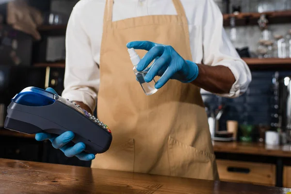 Vista recortada de terminal de pago y antiséptico en manos de barista afroamericano en guantes de látex - foto de stock