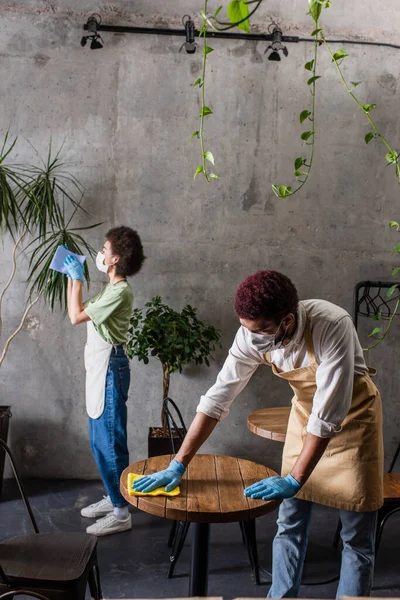 Barista afroamericano en guantes de látex mesa de limpieza cerca de colega en la cafetería — Stock Photo