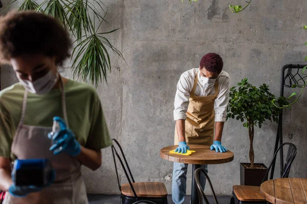 Barista afroamericano in maschera medica tavolo di pulizia vicino sfocato collega — Foto stock