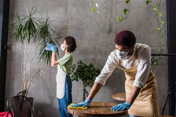 Barista afroamericano in grembiule e mascherina medica tavolo pulizia vicino collega su sfondo sfocato — Foto stock