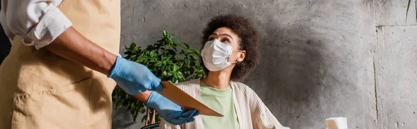 Garçom afro-americano em luvas de látex segurando menu perto do cliente em máscara médica, banner — Fotografia de Stock