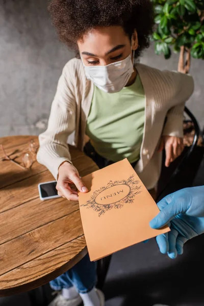 Cliente afroamericano en el menú de celebración máscara médica cerca de camarero en guante de látex en la cafetería - foto de stock