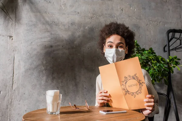 African american woman in medical mask holding menu near smartphone and eyeglasses in cafe — Stock Photo