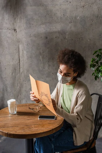 Cliente afroamericano en máscara médica mirando el menú cerca de anteojos y teléfonos inteligentes en la cafetería - foto de stock