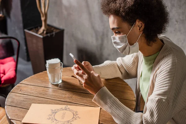 Африканська американка в медичній масці користується смартфоном біля меню в кафе. — стокове фото
