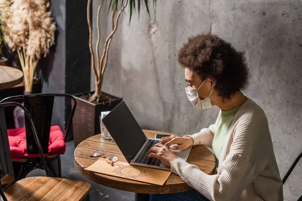 Freelancer afroamericano en máscara médica usando laptop cerca del menú en la cafetería - foto de stock