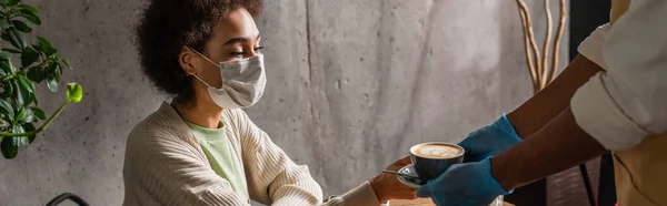 Mujer afroamericana en máscara médica tomando café de camarero en guantes de látex, pancarta - foto de stock