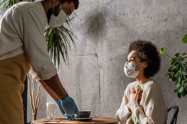 Cliente afroamericano en máscara médica sentado cerca de barista con café en la cafetería - foto de stock