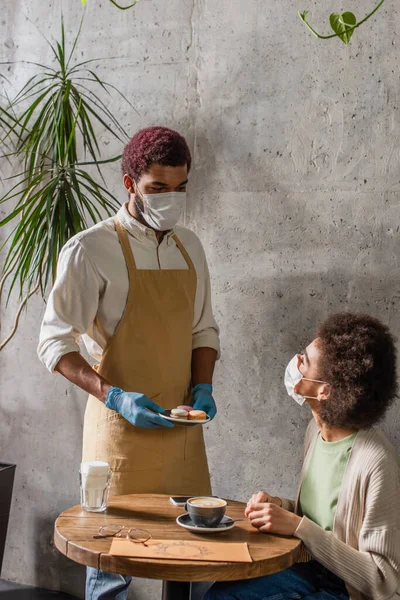 Cameriere afroamericano in guanti di lattice in possesso di amaretti vicino al cliente in maschera medica in caffè — Foto stock